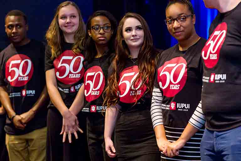 Six IUPUI student volunteers wearing 50th Anniversary T-shirts stand on stage during an event.