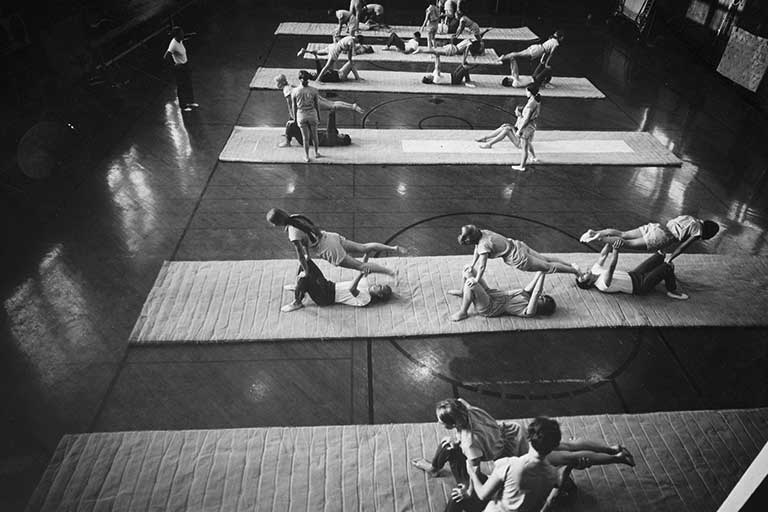 Rows of students sitting on mats practice gymnastics. Black and white photo.
