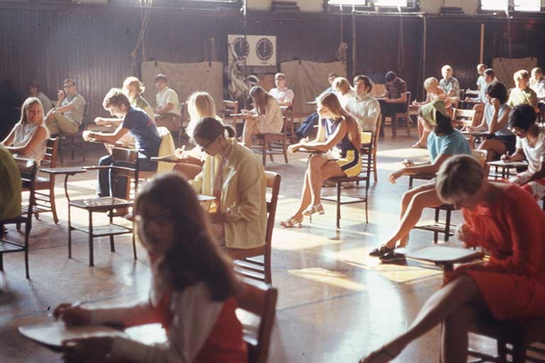 Students sitting at wooden desks take a test in the German House gymnasium, home of the Normal College. Color photo.